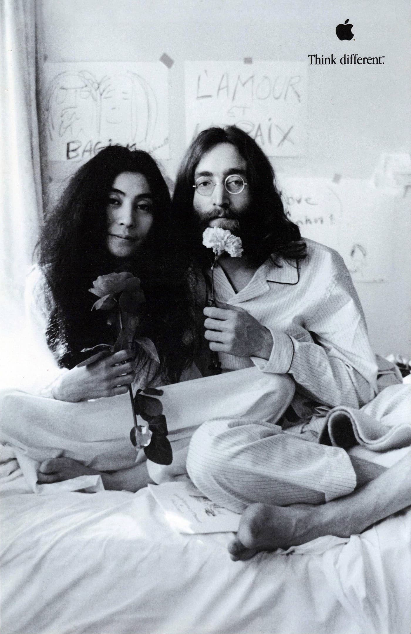 A man and a woman in their pajamas are sitting in bed holding up flowers as a gesture of peace