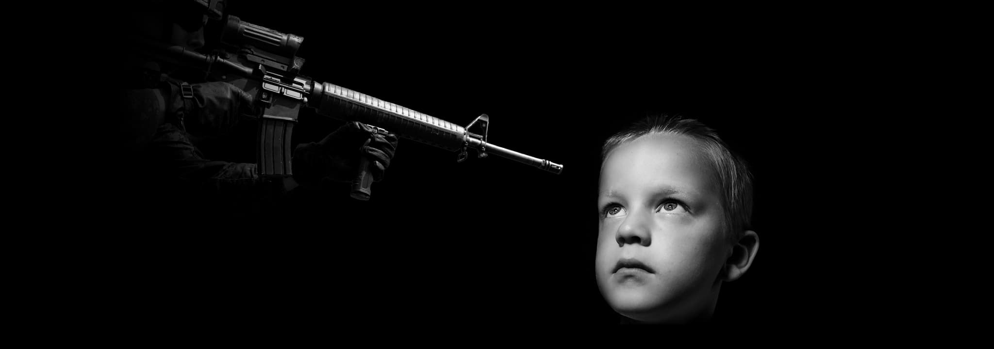 A powerful black-and-white image depicts a young boy looking upwards with a solemn, contemplative expression. On the left, the barrel of an assault rifle held by a gloved hand is pointed, symbolizing the grim reality of gun violence and its impact on children. The dark background heightens the emotional tension, underscoring the unsettling juxtaposition of innocence and the threat of violence in modern society.