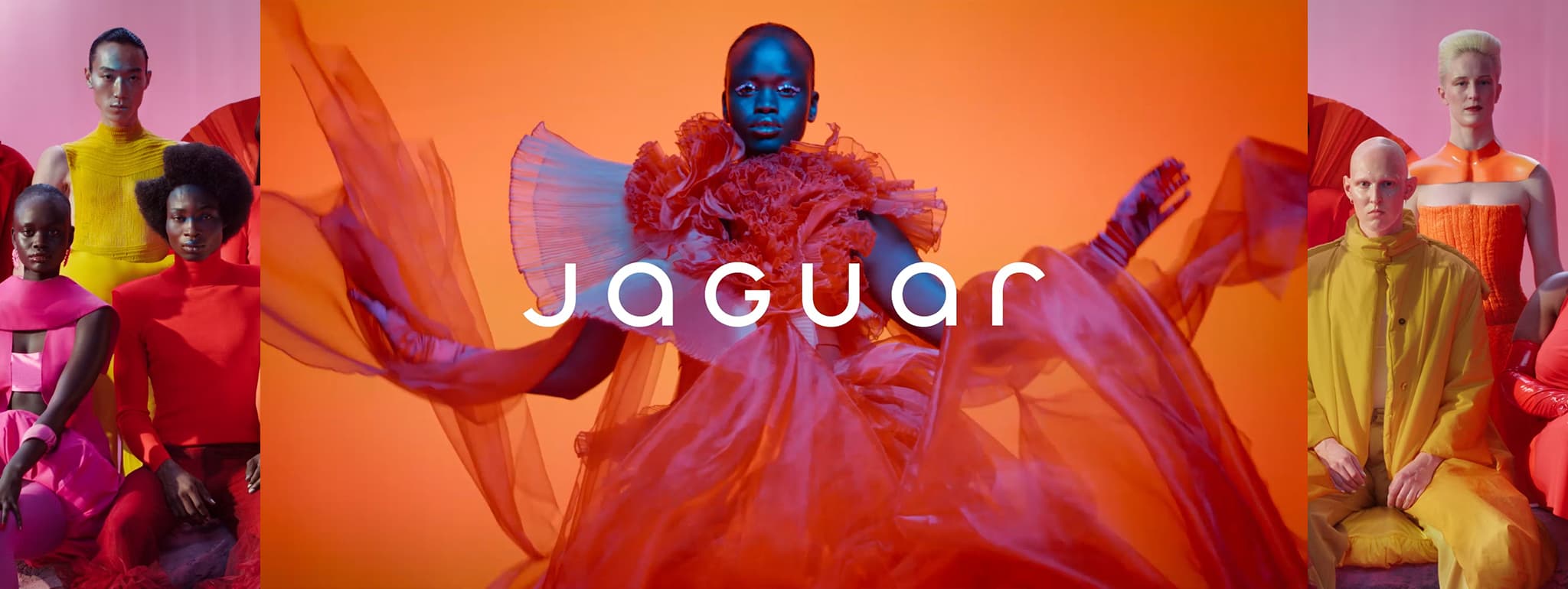 Vibrant artistic composition featuring diverse models in striking, colorful fashion. The central figure is dressed in an elaborate orange-red gown, surrounded by models in bold outfits of pink, red, yellow, and orange tones. The background transitions between shades of orange and pink, with the word ‘JAGUAR’ displayed prominently in the center.