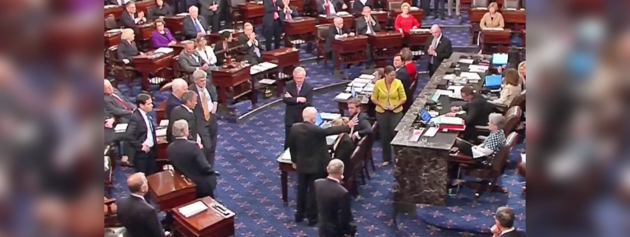 Senator John McCain standing in the center of the U.S. Senate floor, surrounded by colleagues, casting his decisive vote against the Obamacare repeal, while others observe and react.