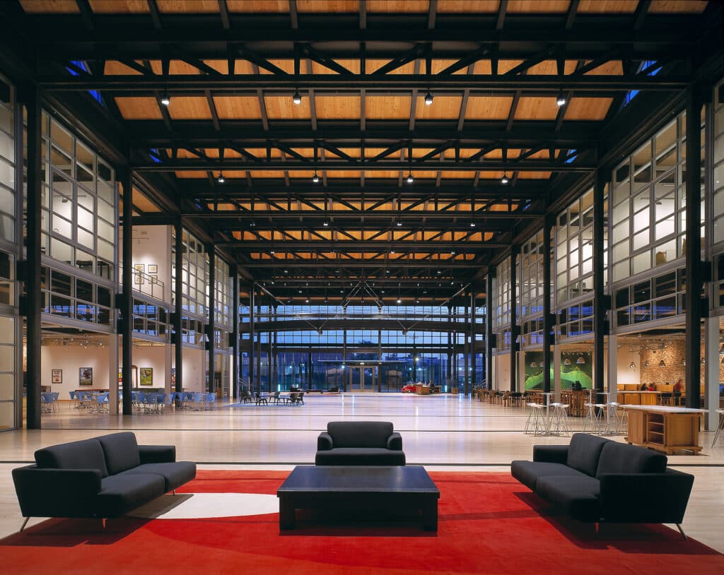 Modern open space with exposed wooden ceiling beams and steel structure. Features floor-to-ceiling windows, polished concrete floors, and a central seating area with black couches arranged on a red carpet. Café-style seating visible along the walls with art displays.