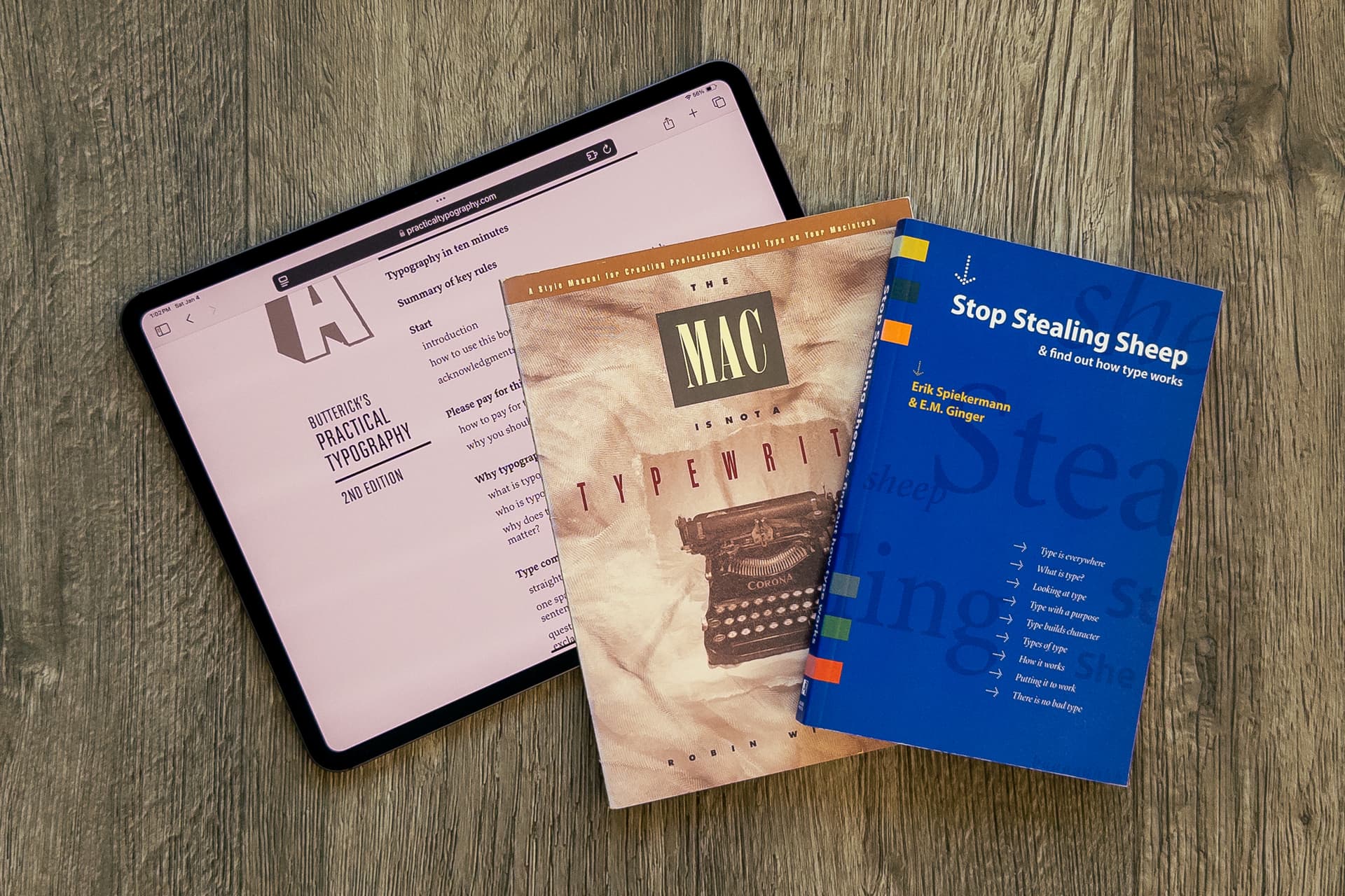 An iPad displaying "Butterick's Practical Typography 2nd Edition" alongside two typography-focused books: "The Mac is not a typewriter" with a vintage typewriter on its cover, and a blue book titled "Stop Stealing Sheep & find out how type works" by Erik Spiekermann and E.M. Ginger. The items are arranged on a weathered wood surface.