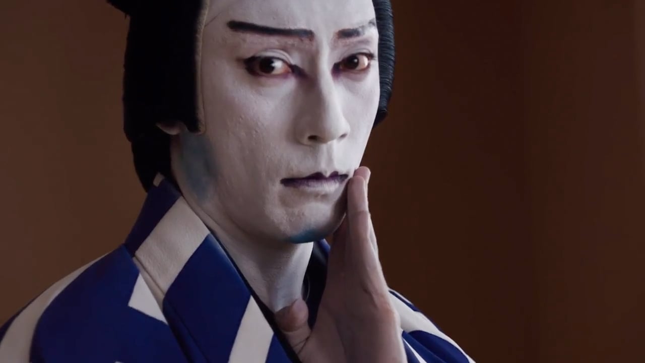 Person in traditional Japanese kabuki makeup and costume, with white face paint, dark eyebrows, and a blue-and-white patterned robe, holding a hand near their cheek in a dramatic pose.